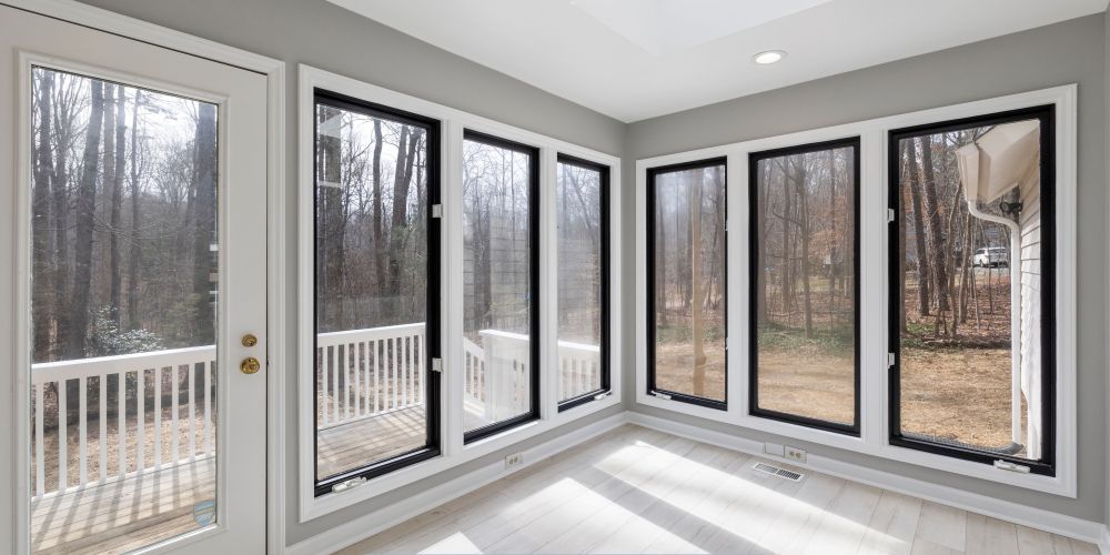 Bright interior sunroom featuring large replacement windows with black frames and energy-efficient glass, allowing natural light and clear outdoor views of a wooded backyard and deck.