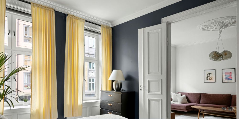 Modern interior bedroom with large white-framed, soft yellow curtains, dark accent wall, and natural light overlooking a city street