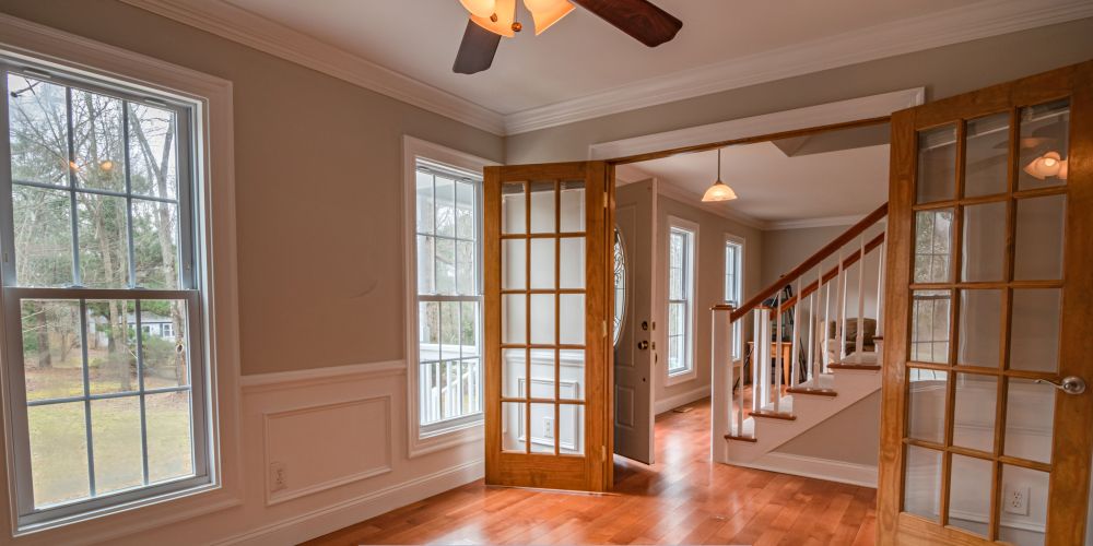 Bright interior room with large double-hung windows, hardwood floors, and wooden French doors leading to an entryway with staircase and natural light