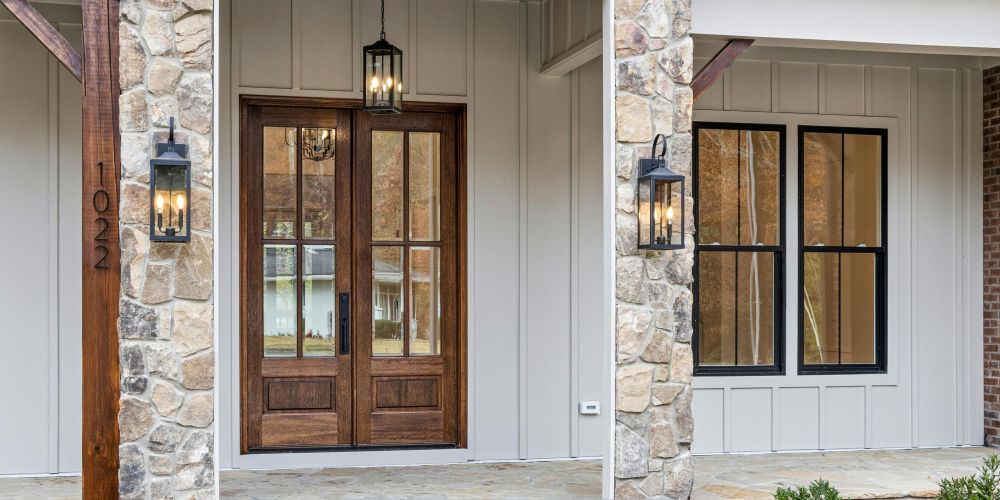 Modern home exterior featuring a wooden double entry door with glass panels, black-framed windows, stone columns, and decorative lantern-style wall lighting on a covered porch